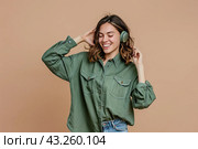 A charming teenage girl puts on wireless headphones, listens to music on a beige cover background. Стоковое AI-изображение, AI-художник Ярославский Максим / Фотобанк Лори