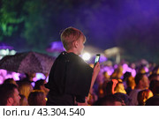 Kaliningrad, Russia - August 20, 2022: Youth is excited during street rock concert. They dance. The girl sits on shoulders of guy and makes photos on smartphone. Bright blue neon lighting.  Back view. Редакционное фото, фотограф YuryKara / Фотобанк Лори