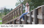 Woman with headphones dangles her legs while sitting on fence in public park. Стоковое видео, видеограф Владимир Григорьев / Фотобанк Лори
