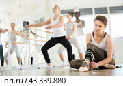 Ballerina ties her pointe shoes on her feet during break between ballet rehearsals. Other participants continue to perform various ballet steps. Стоковое фото, фотограф Яков Филимонов / Фотобанк Лори