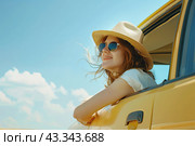 A happy girl in glasses and a straw hat leans out of the window of a yellow car against the blue sky. Стоковое AI-изображение, AI-художник Ярославский Максим / Фотобанк Лори