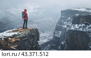 A man in a red jacket with a hood and a backpack stands on the edge of a cliff. There is snow all around, snow-capped mountains and gorges are visible, creating an atmosphere of solitude and severity. The sky is overcast and snowing. The landscape conveys a feeling of coldness and the power of nature, and the person looks determined and ready for new challenges. Стоковое AI-изображение, AI-художник Анна Кривицкая / Фотобанк Лори