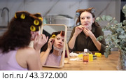 Two carefree adult besties sitting with colorful hair rollers on heads and applying nourishing facial mask to faces, enjoying self care and pampering moment at home together. Стоковое видео, видеограф Яков Филимонов / Фотобанк Лори