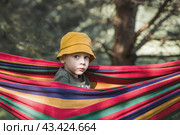 Summer holidays for children in the fresh air.. A cute little boy is having fun with a colorful hammock in the backyard. A child in a hammock. Стоковое фото, фотограф Photojuli / Фотобанк Лори