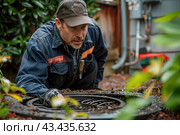 A plumber leaned over a sewer well on the territory of a village house. Septic tank maintenance. Стоковое AI-изображение, AI-художник Коханова Наталья / Фотобанк Лори