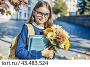A pretty little girl with glasses and a bouquet of autumn flowers goes to school for the first time. A child with a school bag, a book and flowers. A child on a city street. Back to school. Стоковое AI-изображение, AI-художник Photojuli / Фотобанк Лори