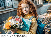 A pretty red-haired girl with glasses and a bouquet of autumn flowers goes to school for the first time. A child with a school bag, a book and flowers. A child on a city street. Back to school. Стоковое AI-изображение, AI-художник Photojuli / Фотобанк Лори