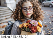 A cute little curly-haired girl with glasses and a bouquet of autumn flowers goes to school for the first time. A child with a school bag, a book and flowers. A child on a city street. Back to school. Стоковое AI-изображение, AI-художник Photojuli / Фотобанк Лори