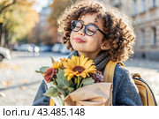 A cute little curly-haired girl with glasses and a bouquet of autumn flowers goes to school for the first time. A child with a school bag, a book and flowers. A child on a city street. Back to school. Стоковое AI-изображение, AI-художник Photojuli / Фотобанк Лори