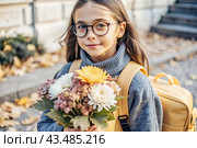 A pretty little girl with glasses and a bouquet of autumn flowers goes to school for the first time. A child with a school bag, a book and flowers. A child on a city street. Back to school. Стоковое AI-изображение, AI-художник Photojuli / Фотобанк Лори
