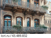 Facade of an ancient building with magnificent decorative elements. The balconies are decorated with elegant openwork wrought iron grilles covered with frost. The windows are decorated with beautiful carved frames and blue curtains. Green plants grow on the balconies, adding beauty and coziness to this winter scene. Стоковое AI-изображение, AI-художник Анна Кривицкая / Фотобанк Лори