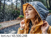 Close-up. A happy young woman in a raincoat enjoys the rain on the background of a green forest, the concept of enjoying a natural leisurely life, environment, lifestyle. Стоковое AI-изображение, AI-художник Photojuli / Фотобанк Лори