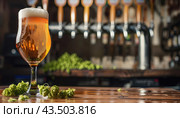 an elegant glass of beer and fresh green hops on a wooden table in a bar against the backdrop of beer pouring taps. copy space. Стоковое AI-изображение, AI-художник Александр Довянский / Фотобанк Лори