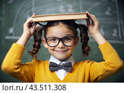 A pretty girl with glasses and books on her head. The child goes to school for the first time. A child in the classroom on the background of a blackboard. Back to school. Стоковое AI-изображение, AI-художник Photojuli / Фотобанк Лори