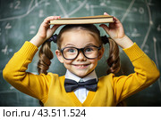 A pretty girl with glasses and books on her head. The child goes to school for the first time. A child in the classroom on the background of a blackboard. Back to school. Стоковое AI-изображение, AI-художник Photojuli / Фотобанк Лори