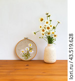 Summer still life with floral handmade embroidery with satin ribbons of daisies flowers in hoops and bouquet of daisies in a vase on wooden table. Crafts and embroidery as a hobby and leisure. Стоковое фото, фотограф Виктория Катьянова / Фотобанк Лори