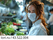 Young female chef in a professional kitchen. She is wearing a white chef's uniform and a medical mask, emphasizing safety precautions and hygiene in the workplace. The woman looks straight into the camera, her hair is neatly tied up, but a few strands are falling out of her hairstyle. In the background you can see the kitchen workspace with various ingredients and herbs, creating an atmosphere of busyness and professionalism. Стоковое AI-изображение, AI-художник Анна Кривицкая / Фотобанк Лори