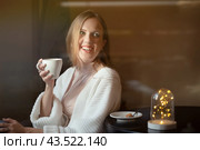 Beautiful happy girl with brown hair is drinking coffee. Attractive young woman in a white sweater with a cup of coffee in her hands. The joyful girl smiles and looks aside. Festive mood. A cozy cafe. Стоковое фото, фотограф Андрей Степанов / Фотобанк Лори