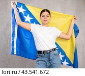 Contented young woman holding Bosnia flag against unicoloured background. Стоковое фото, фотограф Яков Филимонов / Фотобанк Лори