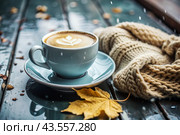 A seasonal drink. Delicious hot cappuccino in a mug with a saucer and a knitted snood, a scarf on a wooden table in the interior of the cafe. It's a rainy autumn day. Стоковое AI-изображение, AI-художник Photojuli / Фотобанк Лори