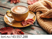 A seasonal drink. Delicious hot cappuccino in a mug with a saucer and a knitted snood, a scarf on a wooden table in the interior of the cafe. It's a rainy autumn day. Стоковое AI-изображение, AI-художник Photojuli / Фотобанк Лори
