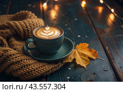 A seasonal drink. Delicious hot cappuccino in a mug with a saucer and a knitted snood, a scarf on a wooden table in the interior of the cafe. It's a rainy autumn day. Стоковое AI-изображение, AI-художник Photojuli / Фотобанк Лори