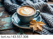 A seasonal drink. Delicious hot cappuccino in a mug with a saucer and a knitted snood, a scarf on a wooden table in the interior of the cafe. It's a rainy autumn day. Стоковое AI-изображение, AI-художник Photojuli / Фотобанк Лори