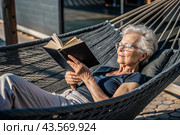 An elderly pensioner, a modern grandmother with glasses, reads a book on vacation in a hammock by the sea in natural light. Стоковое AI-изображение, AI-художник Photojuli / Фотобанк Лори
