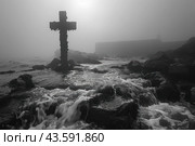 Gloomy seascape in black and white. An old cross stands in the center of the frame, surrounded by seaweed and marine fouling, and partially submerged in water. Water flows rapidly around the cross stones, creating the effect of movement. In the background you can see the foggy coastline with the silhouette of a structure, which adds mystery and drama to the scene. The whole composition creates an atmosphere of privacy, melancholy and antiquity. Стоковое AI-изображение, AI-художник Анна Кривицкая / Фотобанк Лори