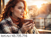 A red-haired, beautiful woman wrapped in a plaid with a mug of hot coffee on a beautiful patio in the rays of the setting sun. The girl inhales the aroma of the drink. Comfort and harmony. Стоковое AI-изображение, AI-художник Photojuli / Фотобанк Лори