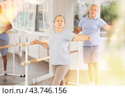 Dancing positive elderly women and men engaged in group class perform exercise near ballet barre, standing in 2nd position of ballet stance in dance studio. Стоковое фото, фотограф Яков Филимонов / Фотобанк Лори