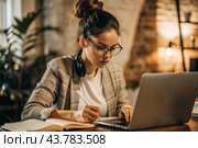 A young female freelance businessman in glasses and headphones is working on a laptop or shopping in an online store. The woman is writing something in a notebook. Стоковое AI-изображение, AI-художник Photojuli / Фотобанк Лори