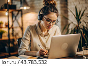 A young female freelance businessman in glasses and headphones is working on a laptop or shopping in an online store. The woman is writing something in a notebook. Стоковое AI-изображение, AI-художник Photojuli / Фотобанк Лори