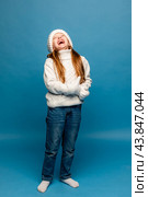 Child girl in a white sweatshirt and knitted hat with pompom on a blue background. The concept of children's emotions, joy and delight. Advertising clothing and sales. Ski resort vacation, copy space. Стоковое фото, фотограф Ирина Михайличенко / Фотобанк Лори