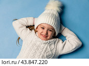 Top view child girl in a white sweatshirt and knitted hat with pompom on a blue background. children emotions, joy and delight. Advertising clothing and sales. Ski resort vacation, copy space. Стоковое фото, фотограф Ирина Михайличенко / Фотобанк Лори