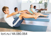 Children doing yoga with group in studio, lying on mat with knee pressed to chest with hands, performing hamstring and low back stretching Ardha Pawanmuktasana. Стоковое фото, фотограф Яков Филимонов / Фотобанк Лори