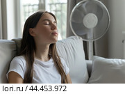 Young woman suffering during a heat heat sitting under air conditioner at home resting on a sofa. Exhausted girl feeling sweaty switching on modern AC system. Hot summer. Fresh clean air in apartment. Стоковое AI-изображение, AI-художник Марина Демидюк / Фотобанк Лори