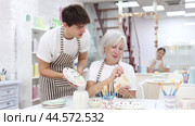 Passionate elderly female teacher offering guidance to interested young guy painting ceramic mug in pottery class. Creative hobby concept. Стоковое видео, видеограф Яков Филимонов / Фотобанк Лори