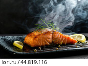 Hot smoked salmon fillet on black plate. Hot lox piece with steamed smoke. Dark background. Salmon steak served lemon, herbs cooking grill barbecue. Grilled red fish dish. Frying trout. Menu, recipe. Стоковое AI-изображение, AI-художник Марина Демидюк / Фотобанк Лори