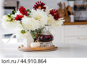 Still-life. White and red dahlia flowers in a vase, a teapot, a pumpkin and a house on a white table in the interior of a home kitchen. A cozy autumn concept. Стоковое фото, фотограф Photojuli / Фотобанк Лори