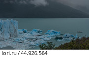 A fragment of a melting glacier. A wall of blue ice over a glacial lake. Melted ice floes float in the water. The tourist boat is sailing. Coastal mountains in clouds, fog. Perito Moreno. El Calafate (2023 год). Стоковое фото, фотограф Вера Ивановна Кубарева / Фотобанк Лори