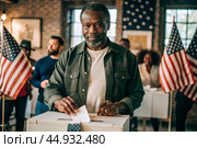 President elections. A black man votes for the new president of the United States of America and puts a form in the ballot box against the background of the American flag. Стоковое AI-изображение, AI-художник Photojuli / Фотобанк Лори
