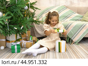 Surprised little smiling girl opening gift box sitting on the floor near the Christmas tree and and rejoices at the contents. Happy girl unwrapping Christmas present looking in box. Стоковое фото, фотограф Ирина Михайличенко / Фотобанк Лори