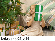 Christmas spirit. Happy little smiling girl opening gift box sitting on the floor near the Christmas tree. Happy girl unwrapping Christmas present looking in box. Holidays and celebrations concept. Стоковое фото, фотограф Ирина Михайличенко / Фотобанк Лори