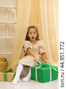 Christmas spirit. Surprised little smiling girl is sitting in the children room in a tent and opening Christmas gifts. Happy girl unwrapping Christmas present box. Holidays and celebrations concept. Стоковое фото, фотограф Ирина Михайличенко / Фотобанк Лори