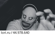 A man in a scary monster mask in a dark attic. Black and white video. The man is wearing a white sweatshirt and a baseball cap on his head. Emotional and dramatic atmosphere. Стоковое видео, видеограф Воловодов Владимир / Фотобанк Лори