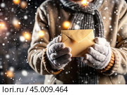 A little girl in winter clothes holding an envelope outside in snowy weather against the background of lights. A letter for Santa Claus. The winter atmosphere of a festive invitation. Стоковое AI-изображение, AI-художник Photojuli / Фотобанк Лори