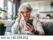 Fraud and deception. A shocked, worried elderly woman looking at her smartphone with unpleasant surprise, experiencing stress, financial problems, money theft. Стоковое AI-изображение, AI-художник Photojuli / Фотобанк Лори