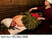 Female with broom do whip steamed man body in russian bathhouse. Male in bath hat lying and sweats on wooden bench in sauna with hot steam. Wellness, self-care, healthy concept. Copy ad text space. Стоковое фото, фотограф Aleksandr Golubev / Фотобанк Лори