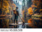 Tourism and sports. A male traveler with a backpack on his back and an oar in his hand is rafting down the river on a sap board against the background of an autumn landscape. Стоковое AI-изображение, AI-художник Photojuli / Фотобанк Лори